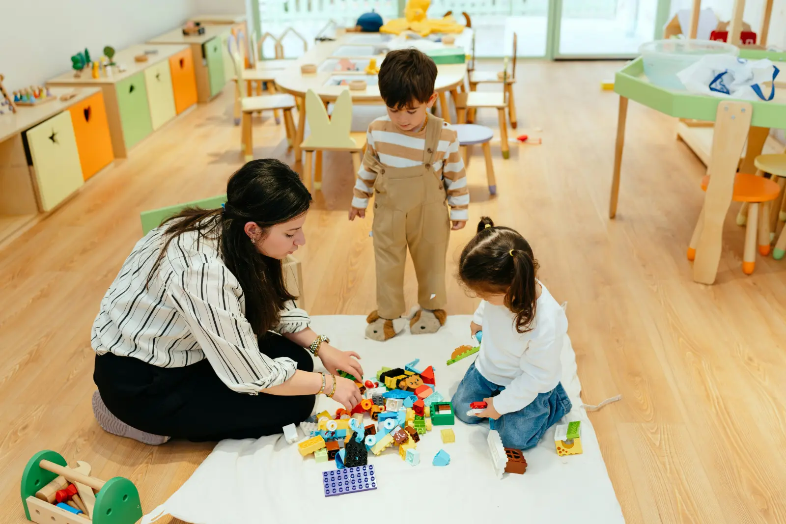 Eine Fachkraft kniet mit zwei kleinen Kindern auf einer Decke und spielt mit bunten Bausteinen.