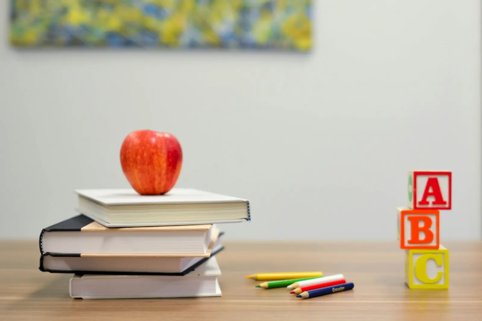 Auf einem Tisch liegen ein Bücherstapel mit einem roten Apfel darauf, bunte Stifte sowie drei Bauklötze mit den Buchstaben A, B und C.