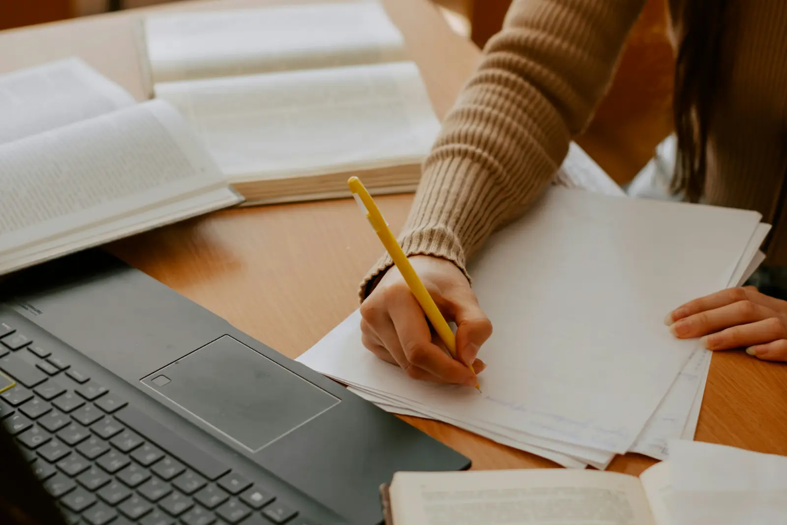 Eine junge Frau schreibt mit Stift auf Papier; um sie herum liegen aufgeschlagene Bücher und ein Laptop auf dem Schreibtisch.